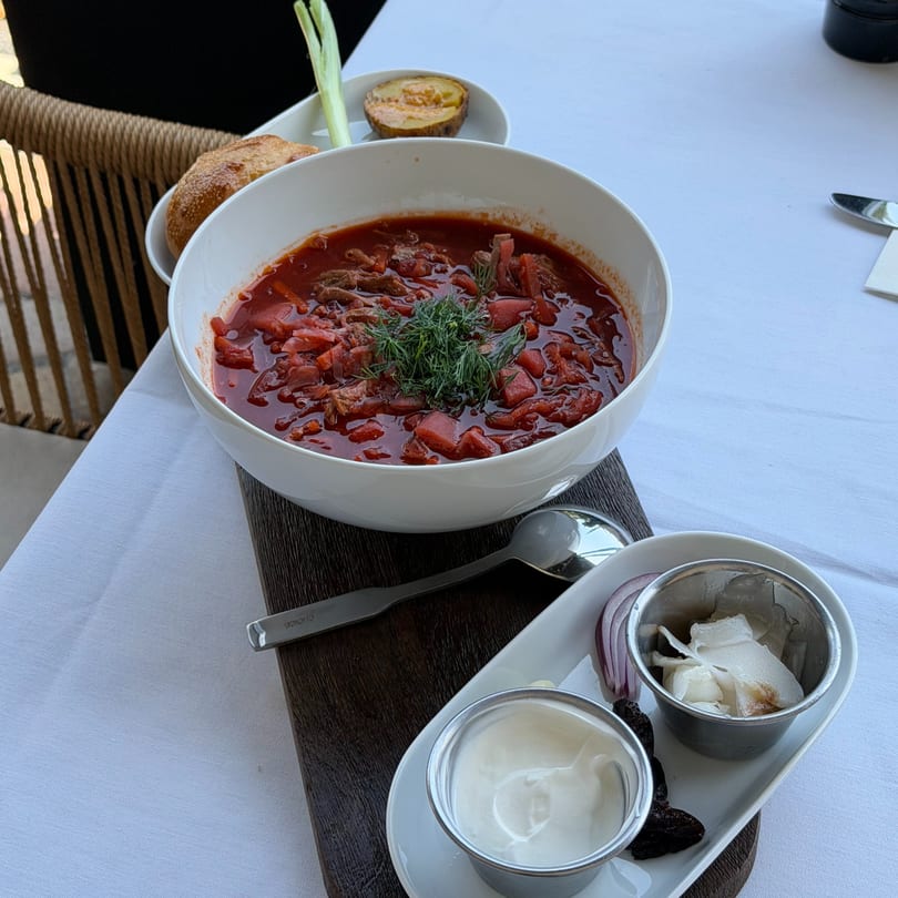 A bowl of borscht with dill, served with bread, potato, sour cream, and garlic on a white tablecloth.
