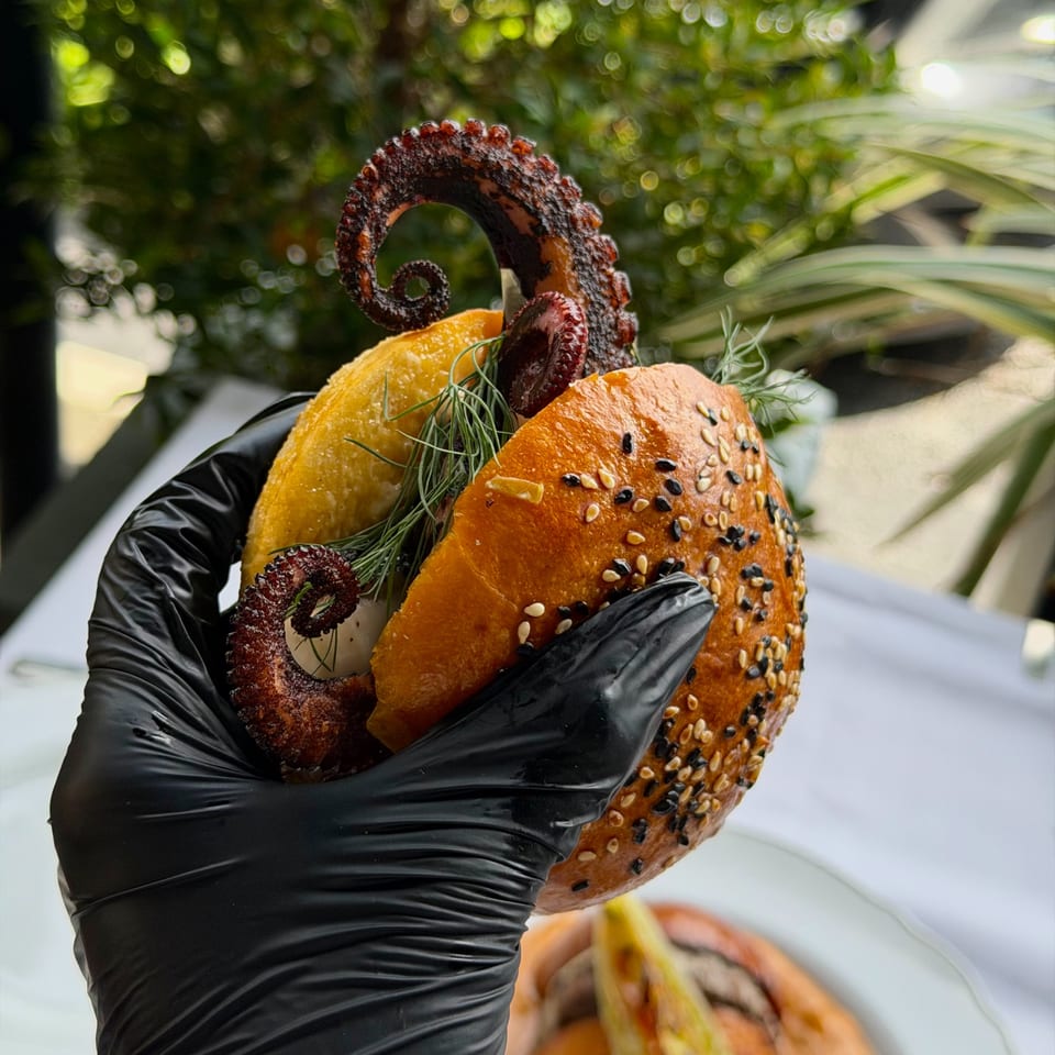 A gloved hand holds an octopus sandwich on a sesame bun with dill, against a green background.