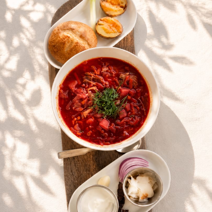 Overhead view of a bowl of borscht with dill, served with bread, potatoes, and condiments.