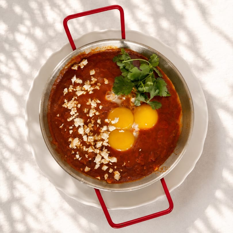 Shakshuka in a pan with three eggs, feta cheese, and cilantro on a white tablecloth.