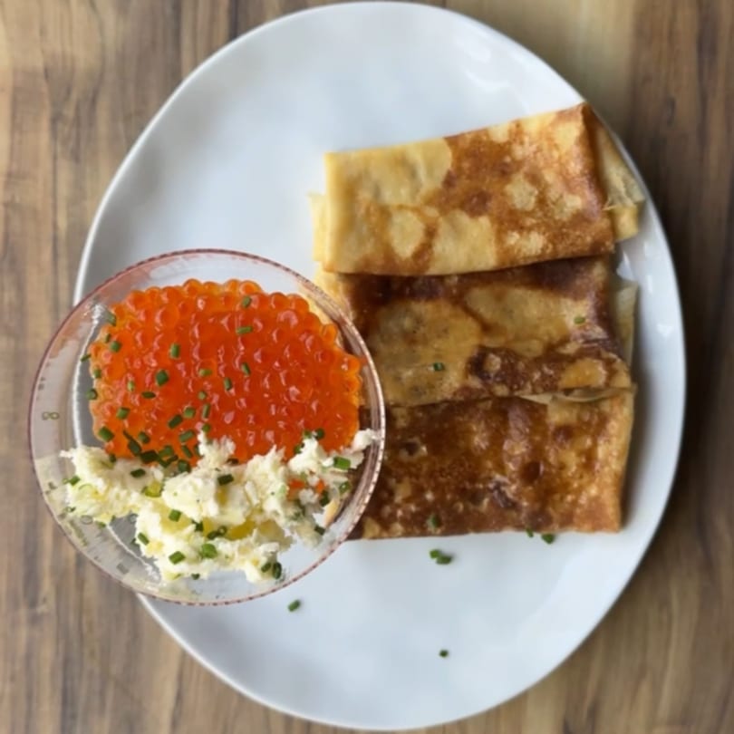 Three golden-brown blinis on a white plate, with a side of red caviar and chopped chives.