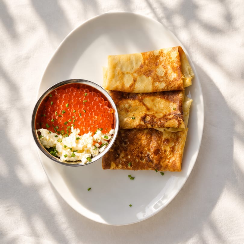 Golden-brown blinis with a side of red caviar and cream cheese, garnished with chives.
